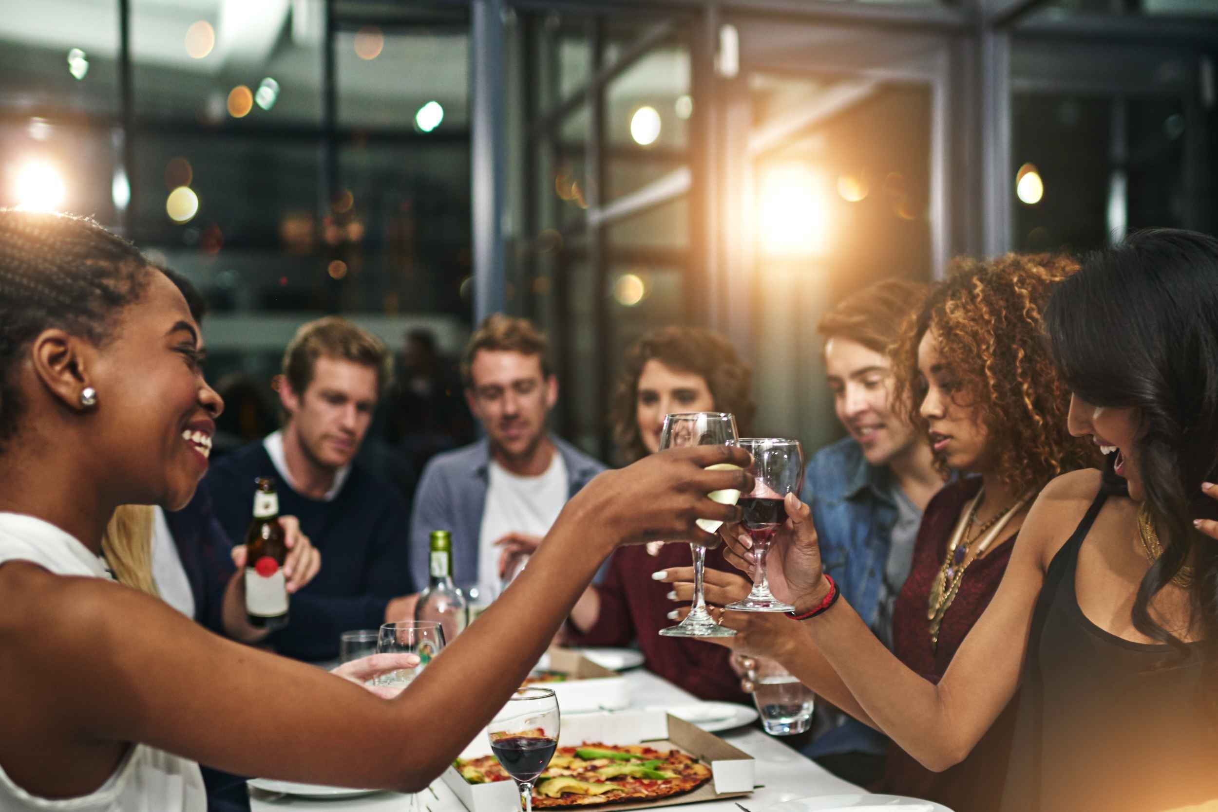 Group of friends toasting with glasses at a dinner table, enjoying food and drinks in a lively setting.