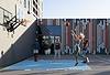 Residents playing basketball on an outdoor court, with one person jumping to shoot the ball while others look on, set against a colorful mural backdrop
