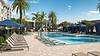 A sunny pool area with lounge chairs, cabanas, and palm trees, featuring blue umbrellas beside the pool.