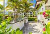Outdoor grilling area surrounded by palm trees, lush greenery, and bright flowers, with a sunny sky in the background.