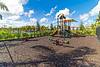 A playground with a slide, climbing structure, and seesaws, surrounded by greenery and a blue sky with clouds.