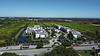 Aerial view of modern buildings near a pond, surrounded by green fields and trees under a clear blue sky.