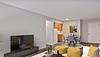 Modern living room with a TV, yellow accent furniture, dining area, and kitchen entrance in a bright, neutral-toned space.