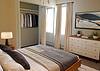 Bright bedroom with a bed, a lamp, a dresser, and a closet with neatly hung clothing. Cozy and modern decor.