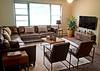Cozy living room with a brown leather sofa, two chairs, a coffee table, a TV, and plants, featuring large windows.