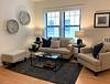 Cozy living room with a beige couch, armchair, coffee table, decorative items, and two wall clocks. Bright and inviting.