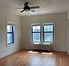 Empty room with hardwood floors, two windows, a ceiling fan, and a radiator. Natural light coming through the windows.