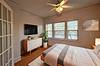 A bright bedroom with a bed, TV, dresser, plant, and large windows allowing natural light. Minimalist decor.