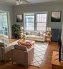 Cozy living room featuring a sofa, two armchairs, coffee table, plants, wall art, and large windows with natural light.