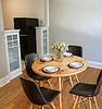A round wooden dining table with four black chairs, set with plates and cutlery, and a vase of flowers in the center.