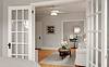 A cozy, light-filled living space with white furniture, a round mirror, and a dining area visible through glass doors.