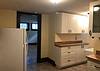 A bright kitchen with white cabinets, a refrigerator, and a doorway leading to a spacious room with wooden flooring.