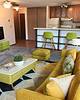 A cozy living room with a yellow sofa, coffee table, green accents, and a kitchenette in the background.