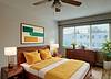 Cozy bedroom with a wooden bed, yellow accents, artwork on the wall, a dresser, and a window with natural light.