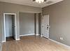 A simple, unfurnished room with beige walls, wooden floor, a ceiling fan, and a door leading outside.