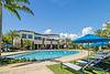 A sunny pool area with lounge chairs and blue umbrellas, surrounded by palm trees and a residential building.