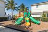 Colorful playground with slides and climbing features, surrounded by palm trees and a fenced area.