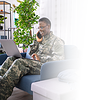 A person in military uniform sits on a couch, using a laptop and talking on a phone, surrounded by greenery.