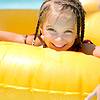 A smiling girl with braids leans on a bright yellow float in a pool, enjoying a sunny day.