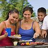 Group of four people enjoying a barbecue, grilling food, and sharing drinks in a sunny outdoor setting.