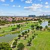 Aerial view of a green park with winding paths, ponds, and residential buildings in the background under a blue sky.