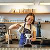A barista pours coffee from a kettle into a filter cone at a stylish café counter, smiling happily.