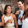 A group of four friends enjoying drinks, smiling and chatting, against a rustic brick backdrop.