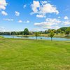 A serene park landscape featuring a grassy area, trees, a calm pond, and a clear blue sky with clouds.