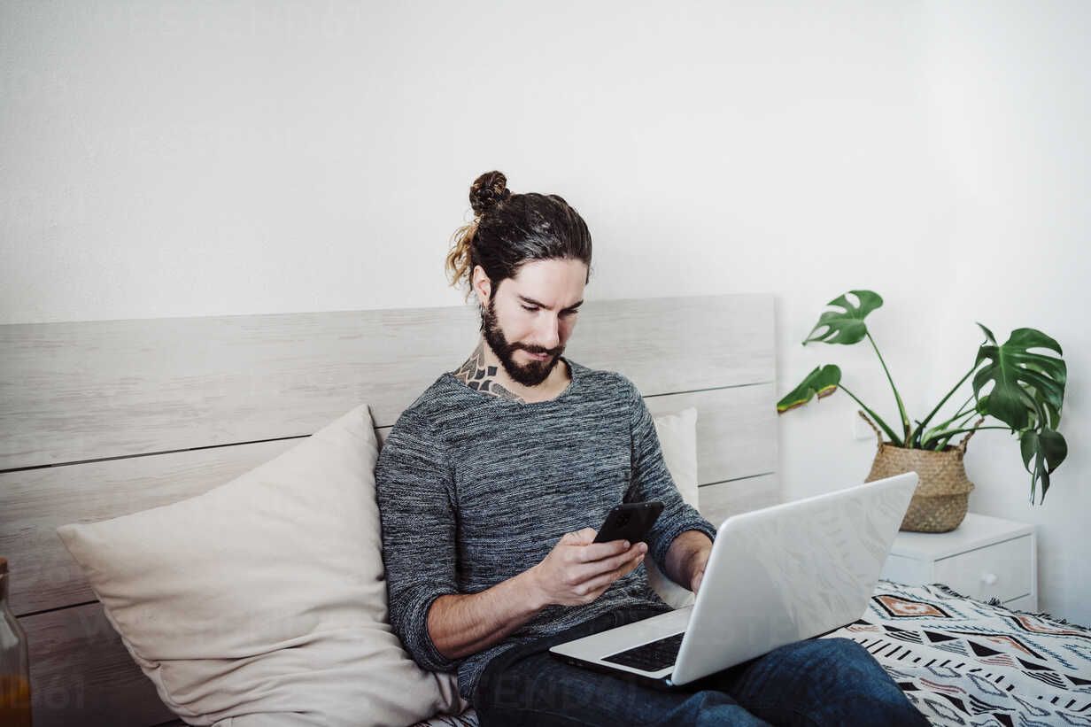 Man using computer and phone while sitting on bed