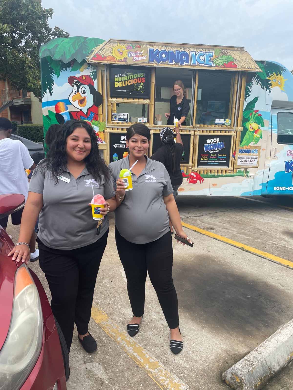 two ladies wearing matching gray shirts standing in front of a Kona Ice truck