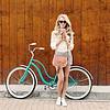 Young blonde woman with long hair with brown purse in sunglasses standing near vintage green bicycle and holding a cup of coffee.